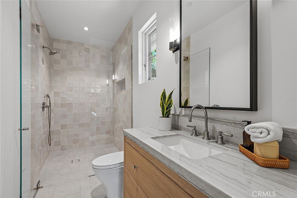 This modern bathroom features a sleek vanity with a light wood finish and a marble countertop, complemented by a large, framed mirror and contemporary lighting. To the left, a spacious walk-in shower with light-toned, textured wall tiles and a glass enclosure adds a luxurious feel. The overall aesthetic is clean, bright, and sophisticated, emphasizing high-end finishes and a minimalist design.
