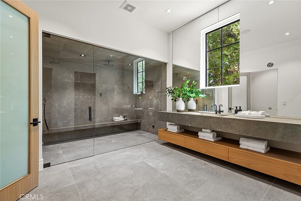 This luxurious primary bathroom features a sleek, modern design with a large walk-in glass-enclosed shower and a floating stone vanity. The space is accented by warm wood cabinetry, a large mirror, and soft recessed lighting, creating a spa-like atmosphere. The neutral color palette and clean lines emphasize a high-end, contemporary aesthetic.