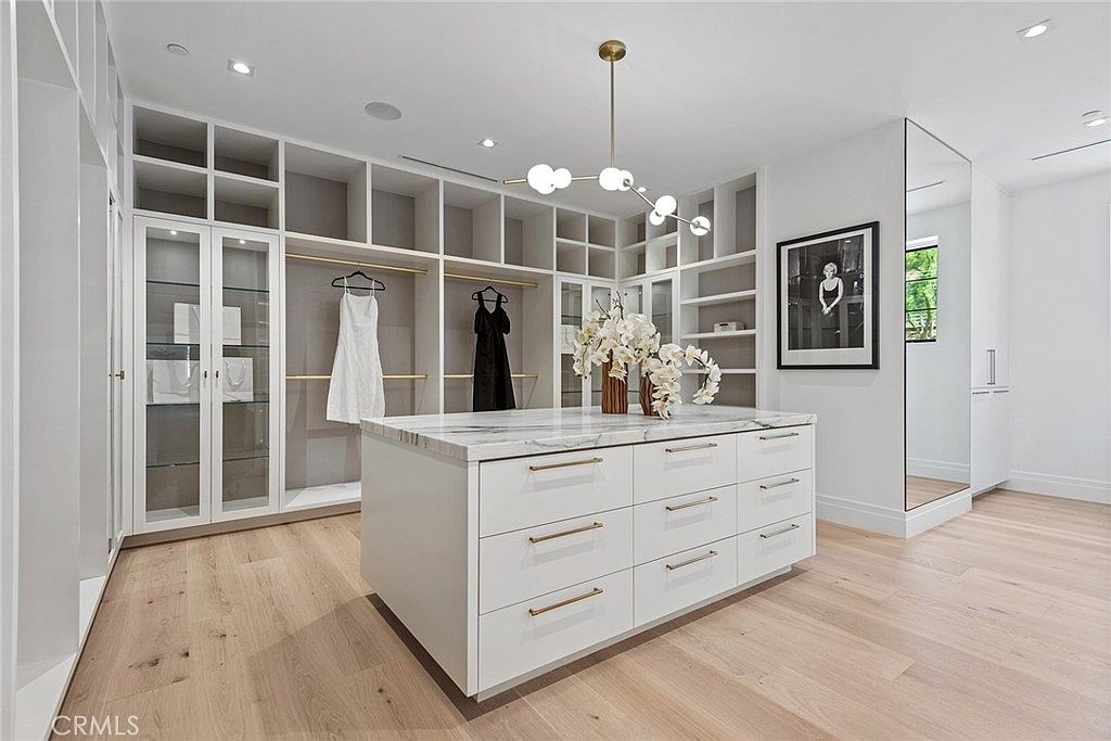 This luxurious, expansive walk-in closet features custom white cabinetry with integrated lighting, providing ample storage and display space. A central marble-topped island with deep drawers serves as a sophisticated focal point, complemented by a modern, multi-globe chandelier. The room is finished with light-toned hardwood flooring and a full-length mirror, creating an airy, high-end boutique atmosphere.