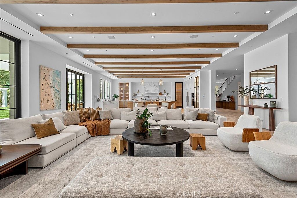 This expansive, open-concept living room features a sophisticated neutral color palette with warm wood ceiling beams that add architectural depth. A massive, plush sectional sofa anchors the space, complemented by a circular coffee table and modern textured accent chairs, all set against a backdrop of high-end finishes and a seamless transition to the kitchen area. The room exudes a luxurious, airy, and inviting atmosphere, perfect for high-end residential living.