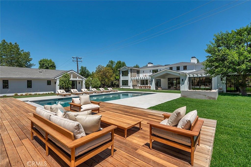 This expansive backyard features a large, rectangular swimming pool as the centerpiece, surrounded by a pristine stone patio and a lush green lawn. In the foreground, a wooden deck is furnished with comfortable, modern outdoor seating, creating an inviting space for relaxation and entertaining. The scene is framed by a luxurious multi-story home with a contemporary design, set against a clear, bright blue sky.