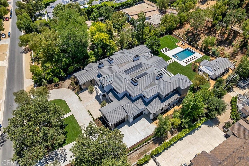 This high-angle aerial view captures a sprawling, modern luxury estate nestled within a lush, tree-lined suburban landscape. The property features a large, multi-gabled grey roof, a spacious concrete driveway, a pristine swimming pool, and a detached guest house, all set against a backdrop of rolling hills. The overall impression is one of exclusivity, privacy, and high-end architectural design.