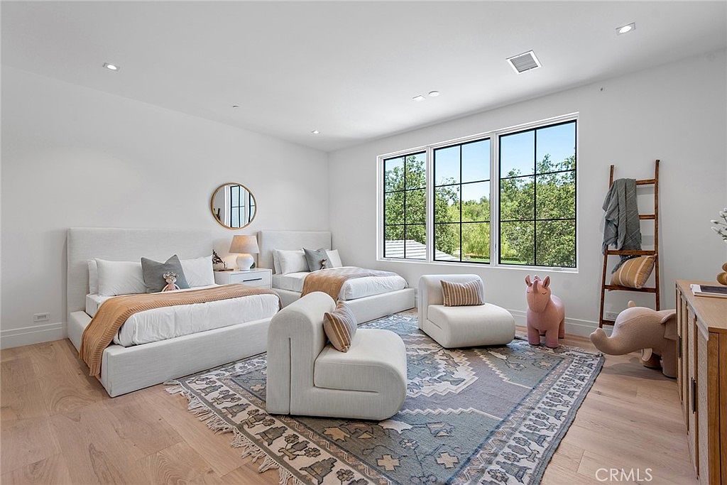 This bright and airy bedroom features two twin beds with neutral-toned bedding, a large patterned area rug, and two modern, low-profile lounge chairs. Large windows with black frames allow for ample natural light and offer a view of the lush greenery outside, while a decorative wooden ladder and whimsical animal-shaped ottomans add a playful, stylish touch to the space.
