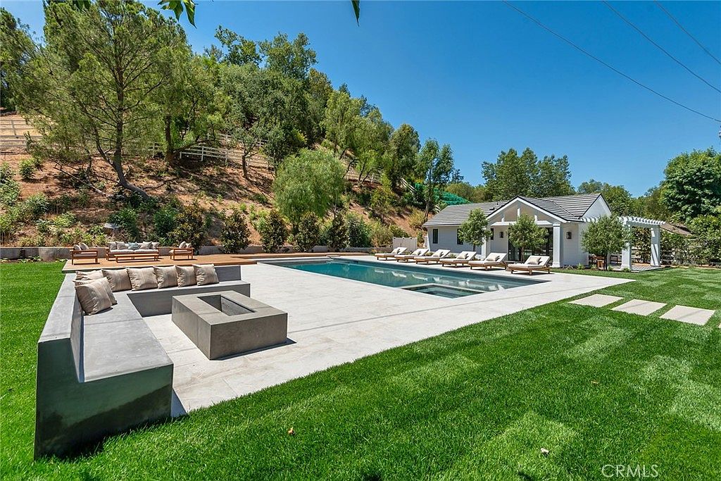 This expansive backyard features a sleek, rectangular swimming pool with an integrated spa, surrounded by a spacious light-colored stone patio. A modern concrete fire pit with built-in bench seating provides a cozy lounge area, while numerous chaise lounges are arranged along the pool deck. The property is set against a lush, tree-covered hillside, creating a serene and private outdoor oasis.