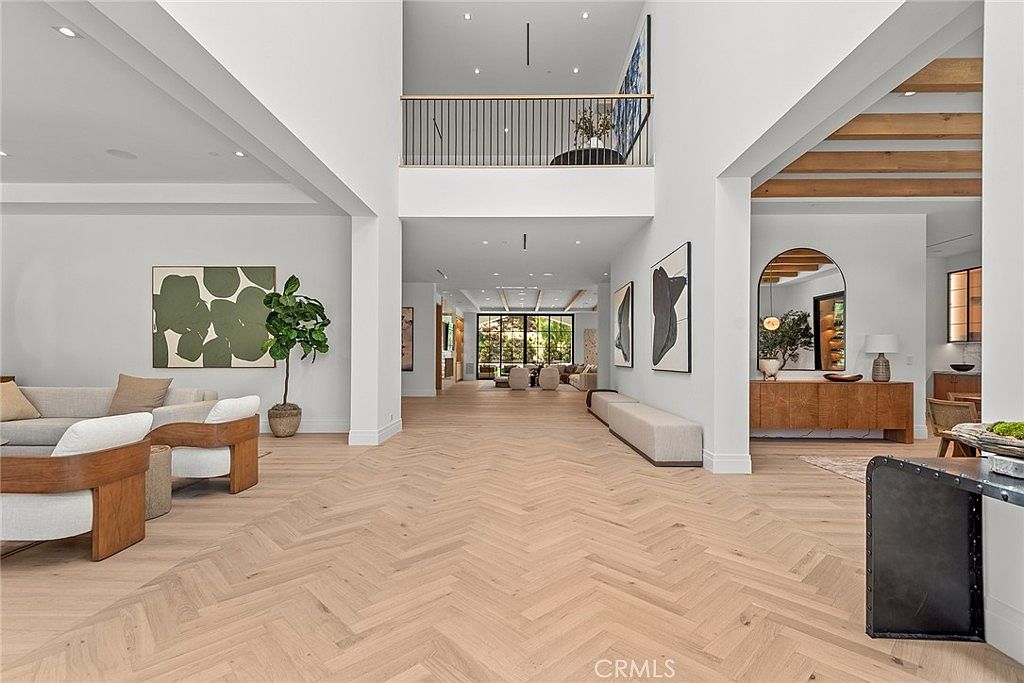This expansive, light-filled foyer showcases a grand open-concept design with high ceilings and elegant light oak herringbone flooring. The space seamlessly transitions from a comfortable living area into a deep, gallery-like hallway that leads toward a bright rear living space, accented by modern art and minimalist decor. The architectural style is sophisticated and airy, emphasizing a sense of luxury and openness throughout the home.