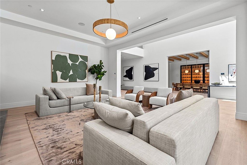 This spacious, modern living room features high ceilings, light hardwood flooring, and a neutral color palette that creates an airy and sophisticated atmosphere. The room is anchored by a large, plush sectional sofa and a contemporary area rug, complemented by a striking drum pendant light and abstract wall art. The open-concept layout provides a clear view into the adjacent dining area, emphasizing the home's expansive and luxurious design.