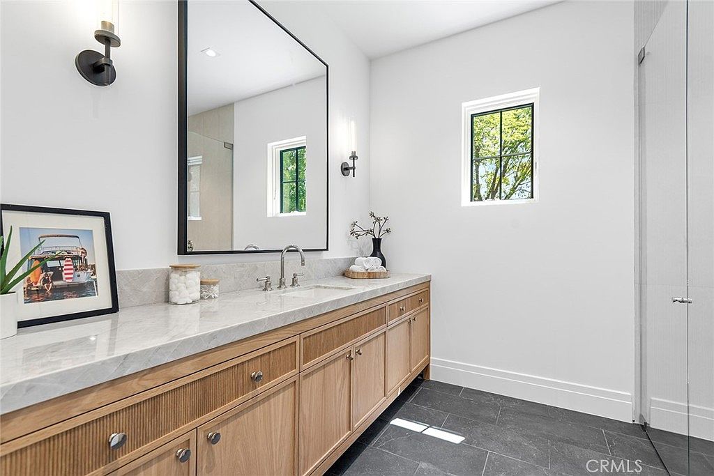 This elegant primary bathroom features a light wood vanity with a marble countertop and a large, sleek rectangular mirror. The space is bright and airy, complemented by dark slate-style floor tiles and a minimalist aesthetic. A framed picture adds a personal touch to the clean, modern design.