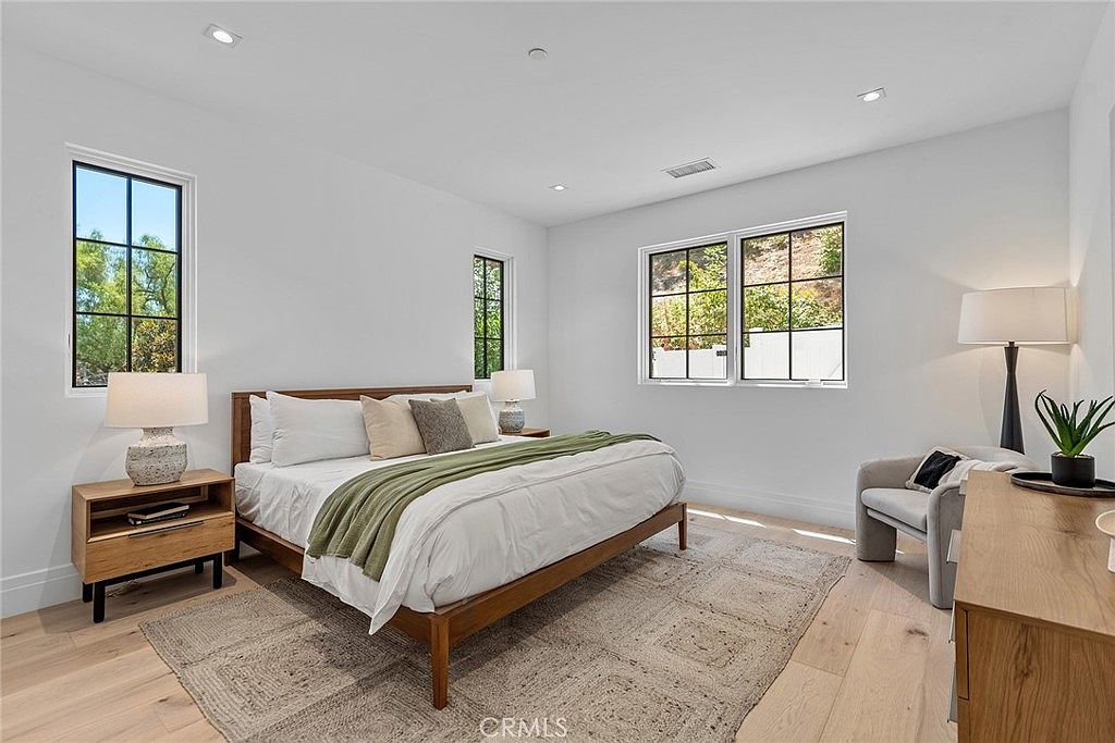 This bright and airy primary bedroom features a modern aesthetic with light wood flooring, crisp white walls, and a large bed dressed in neutral linens with a green throw blanket. The room is accented by two wooden nightstands with matching lamps, a cozy armchair in the corner, and a wooden dresser, all bathed in natural light from multiple windows. The overall impression is one of serene, contemporary comfort and clean design.