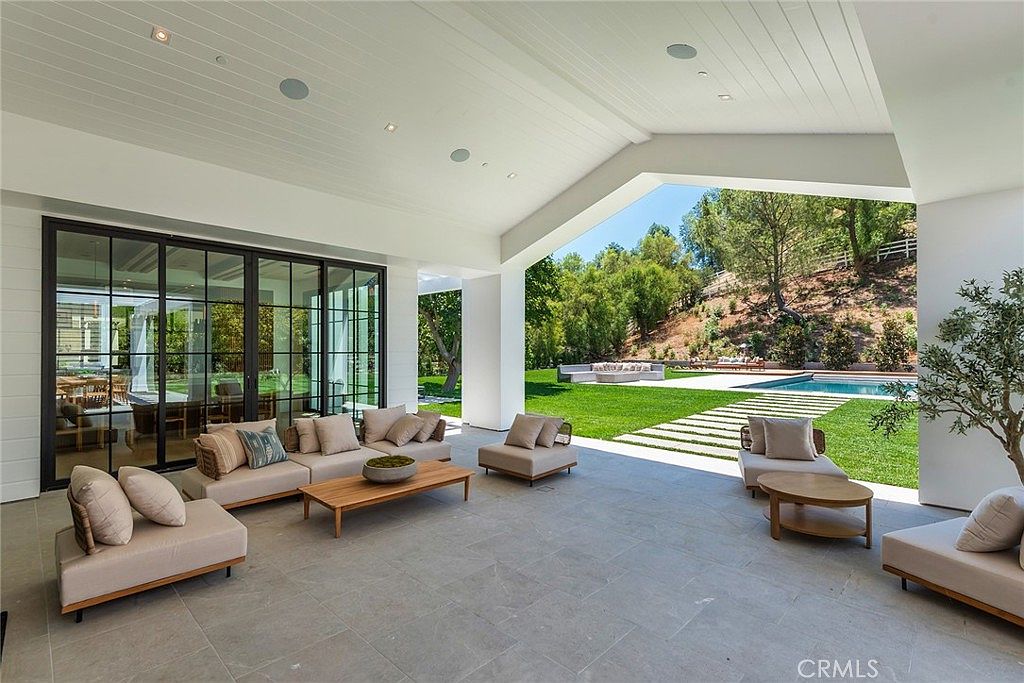 This expansive, covered outdoor living space features a modern, minimalist design with light-toned stone flooring and a high, vaulted ceiling. The area is furnished with comfortable, neutral-colored lounge seating and a wooden coffee table, seamlessly transitioning to a lush backyard with a swimming pool. The open-air layout provides a bright, airy atmosphere perfect for luxury entertaining and indoor-outdoor living.