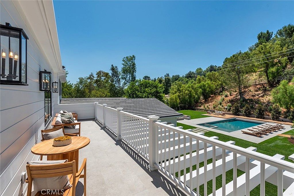 This elevated balcony offers a serene outdoor living space with light-toned tile flooring, comfortable seating, and a wooden bistro set. The white railing provides a clear view of the lush backyard, which features a sparkling swimming pool, manicured lawn, and a scenic hillside backdrop. The bright, sunny atmosphere creates an inviting and luxurious feel, perfect for relaxation or entertaining.