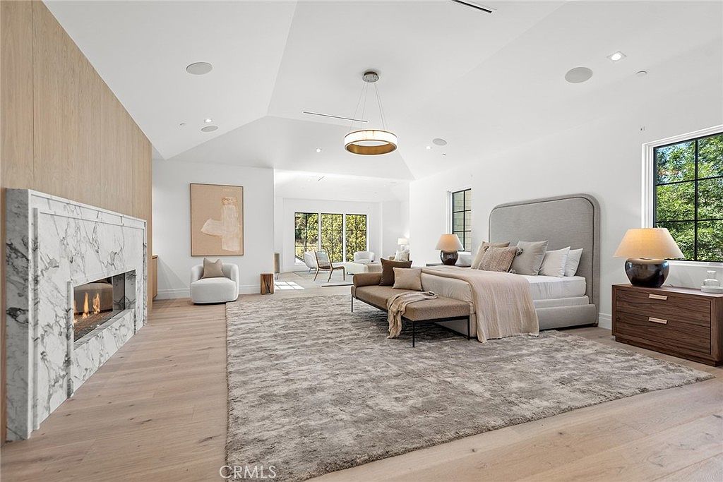 This expansive primary bedroom features a sophisticated, neutral color palette with high vaulted ceilings and light hardwood flooring. A prominent marble-clad fireplace serves as a focal point, while a plush area rug anchors the large bed and seating area, creating a luxurious and serene atmosphere. The room is bathed in natural light from large windows, offering a seamless transition to an adjacent sitting area.