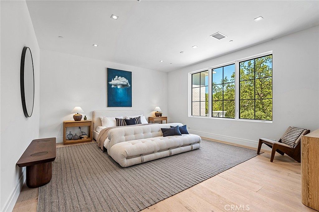 This spacious primary bedroom features a contemporary design with light hardwood flooring, a large area rug, and a neutral color palette. The room is anchored by a plush, tufted bed frame and a matching bench, complemented by minimalist nightstands and a striking piece of abstract wall art. Large windows allow for ample natural light, creating an airy and serene atmosphere.