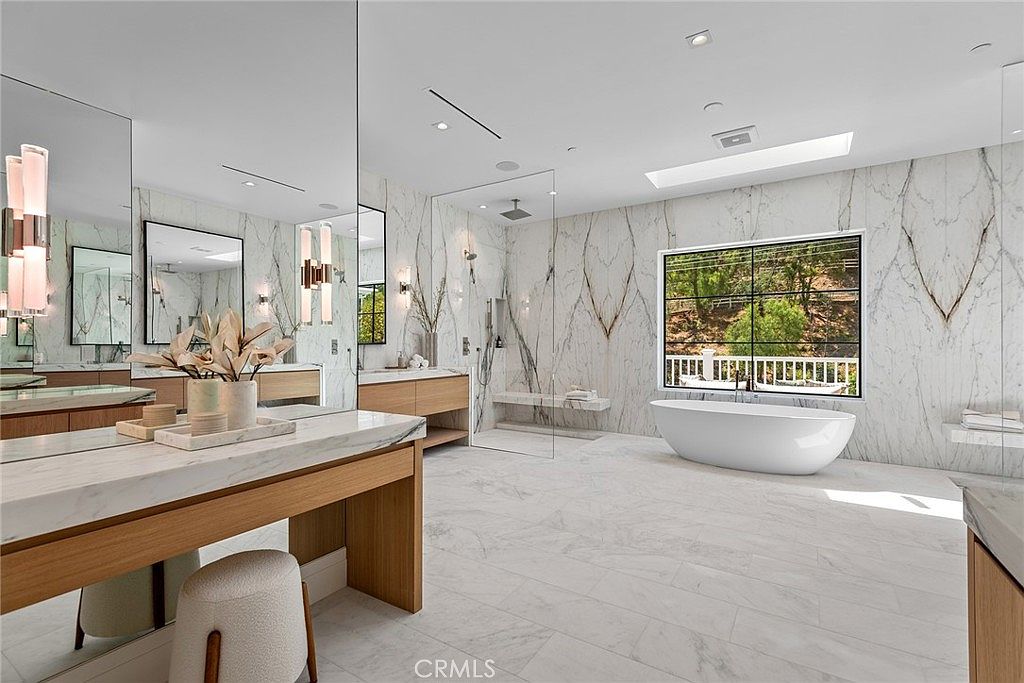 This luxurious primary bathroom features expansive marble wall cladding, a sleek freestanding soaking tub, and a large walk-in glass shower with a built-in bench. The space is anchored by a double vanity with warm wood cabinetry and modern lighting, creating a bright, spa-like atmosphere. A large window provides natural light and a serene view of the surrounding greenery.