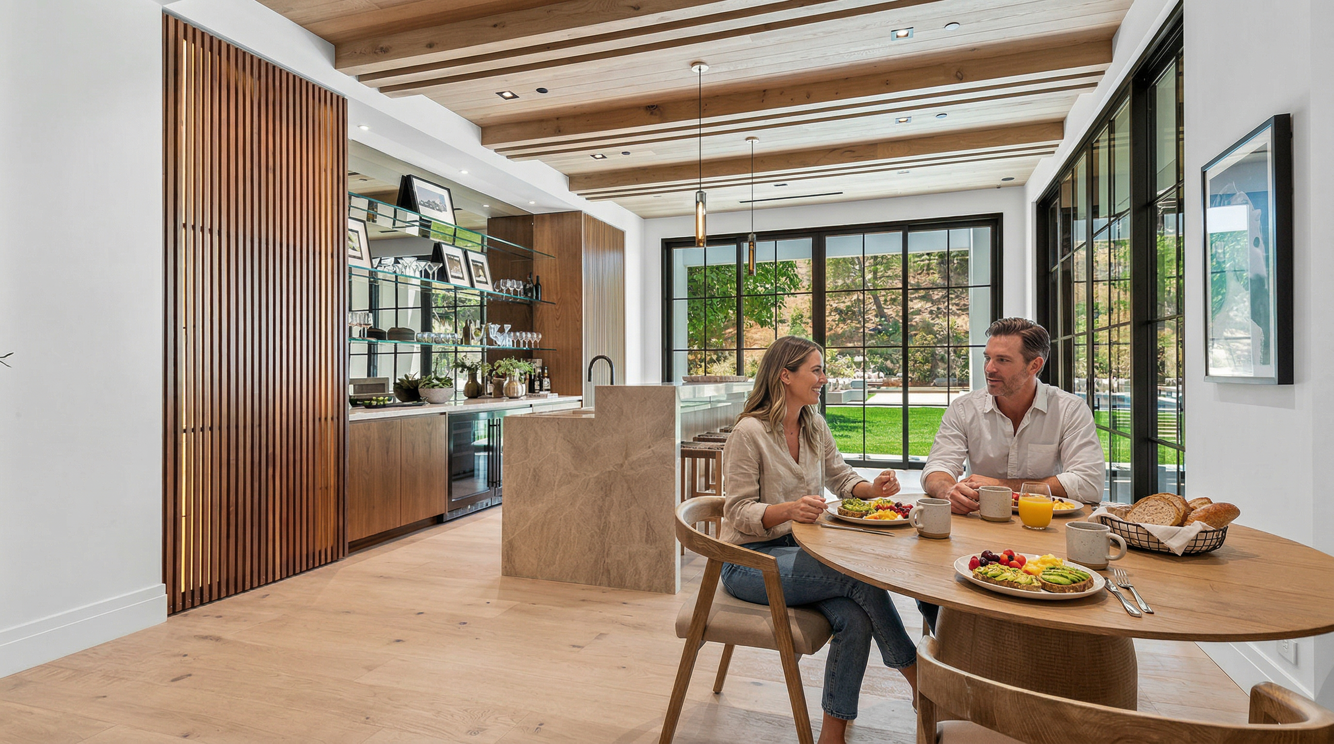 This sophisticated, open-concept kitchen and dining area features a stunning blend of natural wood textures, including a slatted wall and exposed ceiling beams. A large stone-topped island serves as a focal point, complemented by a sleek bar area with glass shelving and a wine fridge. The space is bathed in natural light from floor-to-ceiling black-framed windows that offer a seamless transition to the outdoor landscape.