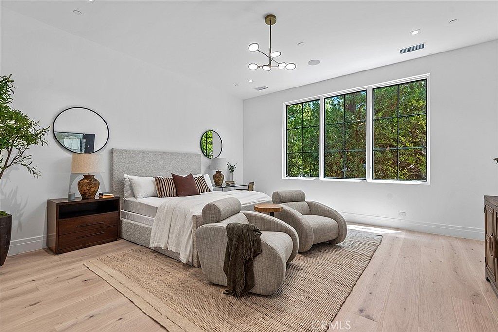 This spacious primary bedroom features a contemporary design with light hardwood floors, a large upholstered bed, and two modern lounge chairs positioned on a textured area rug. Large windows with black frames allow for ample natural light, while a minimalist chandelier adds a touch of elegance to the high ceiling. The room is decorated with circular wall mirrors and warm-toned accents, creating a serene and sophisticated atmosphere.