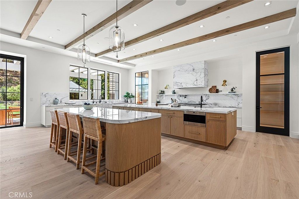 This expansive, high-end kitchen features a large central island with woven bar stools and a marble countertop, complemented by warm wood cabinetry and light hardwood flooring. The space is defined by elegant ceiling beams, modern pendant lighting, and a striking marble backsplash that extends to the ceiling above the range. A large glass-paned door on the right and floor-to-ceiling windows on the left provide a bright, airy atmosphere with a seamless connection to the outdoors.
