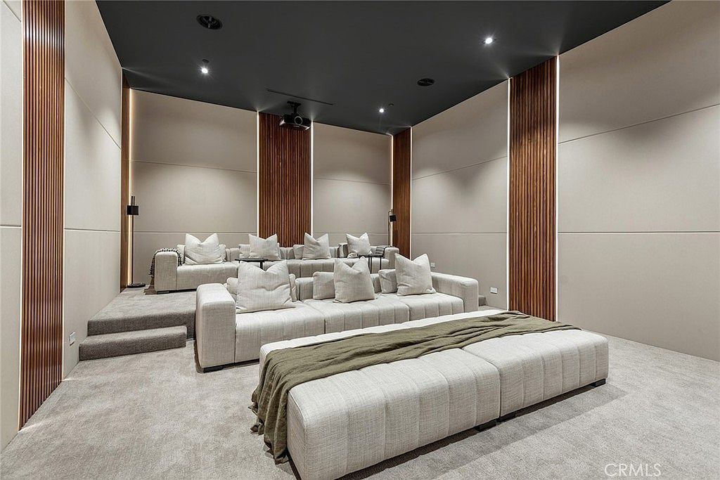This image showcases a luxurious, tiered home theater room designed for ultimate comfort and cinematic immersion. The space features plush, light-colored sectional seating arranged on two levels, accented by warm wood-slat wall panels with integrated vertical lighting. The dark ceiling and neutral color palette create a sophisticated, cozy atmosphere perfect for private screenings.