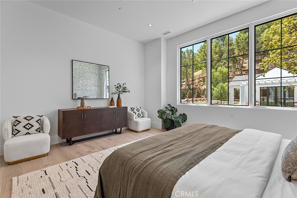 This bright and airy primary bedroom features a neutral color palette, light wood flooring, and a large window that floods the space with natural light. The room is furnished with a comfortable bed, a sleek dark wood dresser, and two stylish accent chairs, creating a serene and sophisticated atmosphere. The perspective is from the foot of the bed, looking toward the wall with the dresser and window, highlighting the clean lines and modern design.