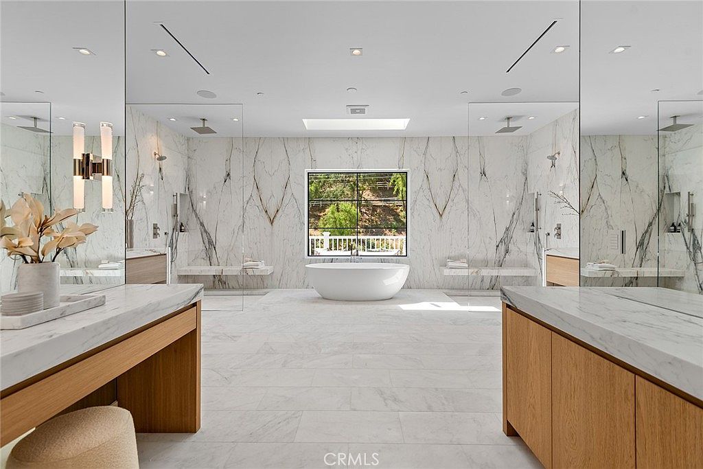 This luxurious primary bathroom features a symmetrical design with dual vanities, expansive marble wall cladding, and a central freestanding soaking tub positioned beneath a window. The space is defined by its clean, modern aesthetic, high-end finishes, and a spacious walk-in shower area enclosed by glass panels. The perspective is centered, emphasizing the grand scale and sophisticated, spa-like atmosphere of the room.