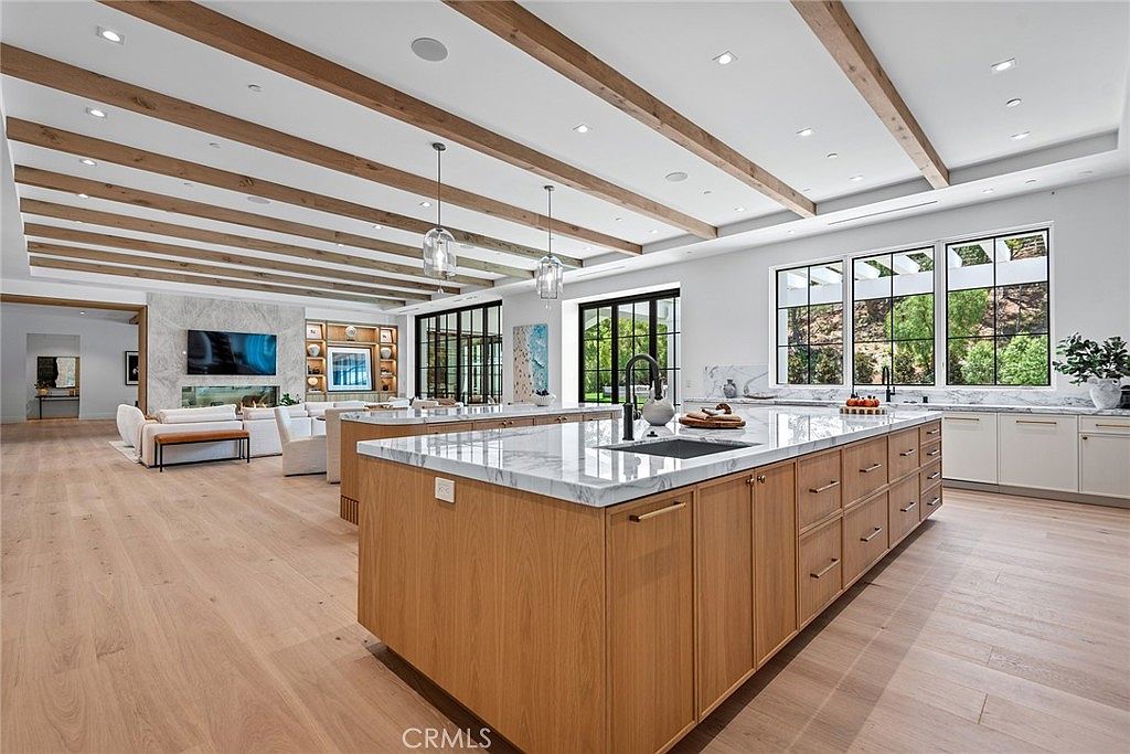 This expansive, open-concept kitchen features a massive central island with a waterfall marble countertop and warm wood cabinetry, creating a sophisticated and inviting atmosphere. The room is highlighted by a striking coffered ceiling with exposed wooden beams and modern pendant lighting, seamlessly flowing into a bright living area with a fireplace. Large windows flood the space with natural light, emphasizing the high-end finishes and spacious, airy design perfect for modern living.