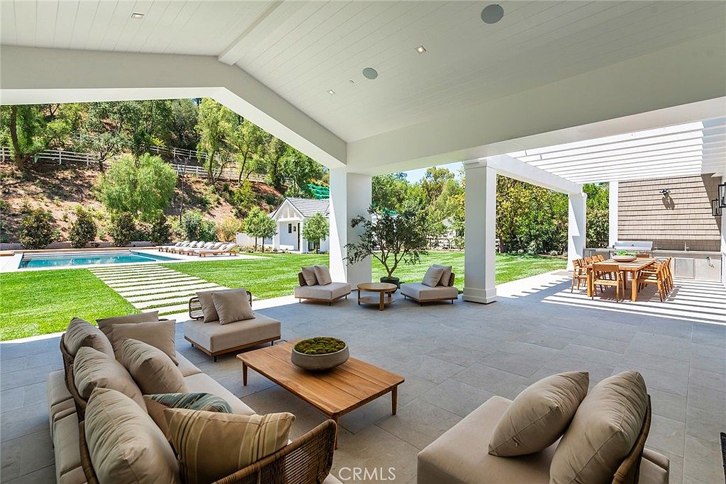 This expansive covered patio offers a seamless transition between indoor and outdoor living, featuring high-end lounge furniture and a central wooden coffee table. The space overlooks a pristine backyard with a sparkling swimming pool, manicured lawn, and a separate dining area under a pergola. The architectural design emphasizes open-air luxury, perfect for entertaining in a serene, sun-drenched setting.