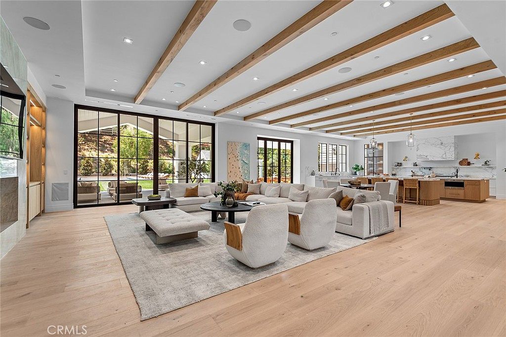 This expansive, open-concept living room features high ceilings with decorative wood beams and light-toned hardwood flooring throughout. A large, plush sectional sofa and two modern armchairs are arranged around a circular coffee table, creating a comfortable seating area that flows seamlessly into a gourmet kitchen in the background. Massive floor-to-ceiling glass doors provide an abundance of natural light and a clear view of the outdoor patio, emphasizing a bright, airy, and luxurious atmosphere.