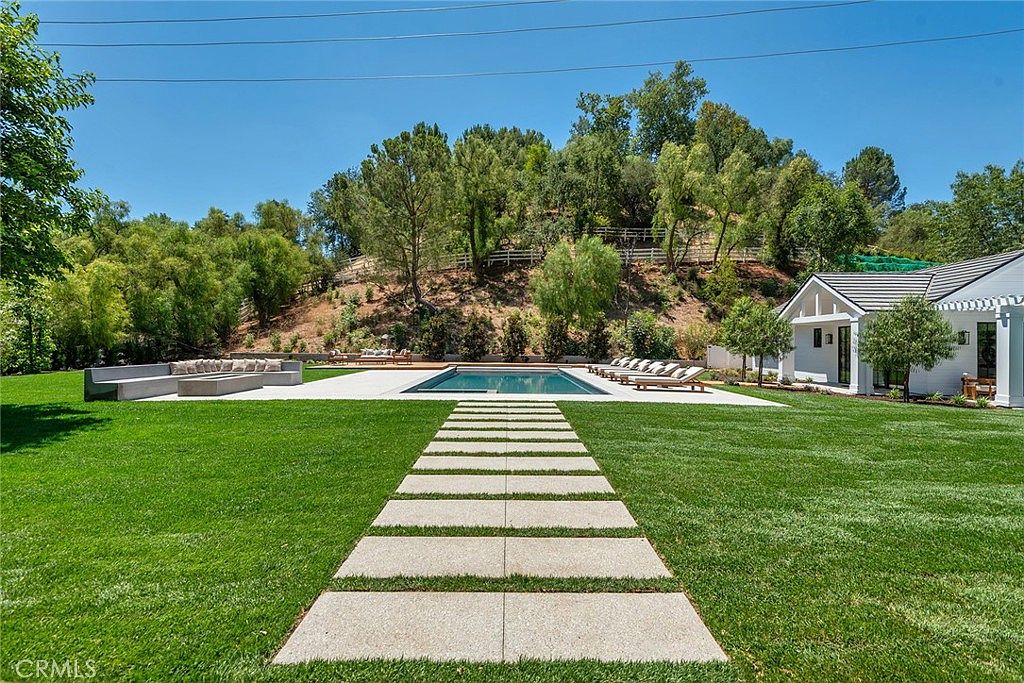 This expansive backyard features a pristine rectangular swimming pool as the central focal point, complemented by a modern stone paver walkway leading through a lush, manicured lawn. To the left, a large contemporary outdoor sectional sofa provides ample seating, while a white-trimmed structure with a dark roof sits to the right, offering a sophisticated architectural contrast. The scene is framed by mature trees and a gentle hillside, creating a serene and luxurious private retreat.