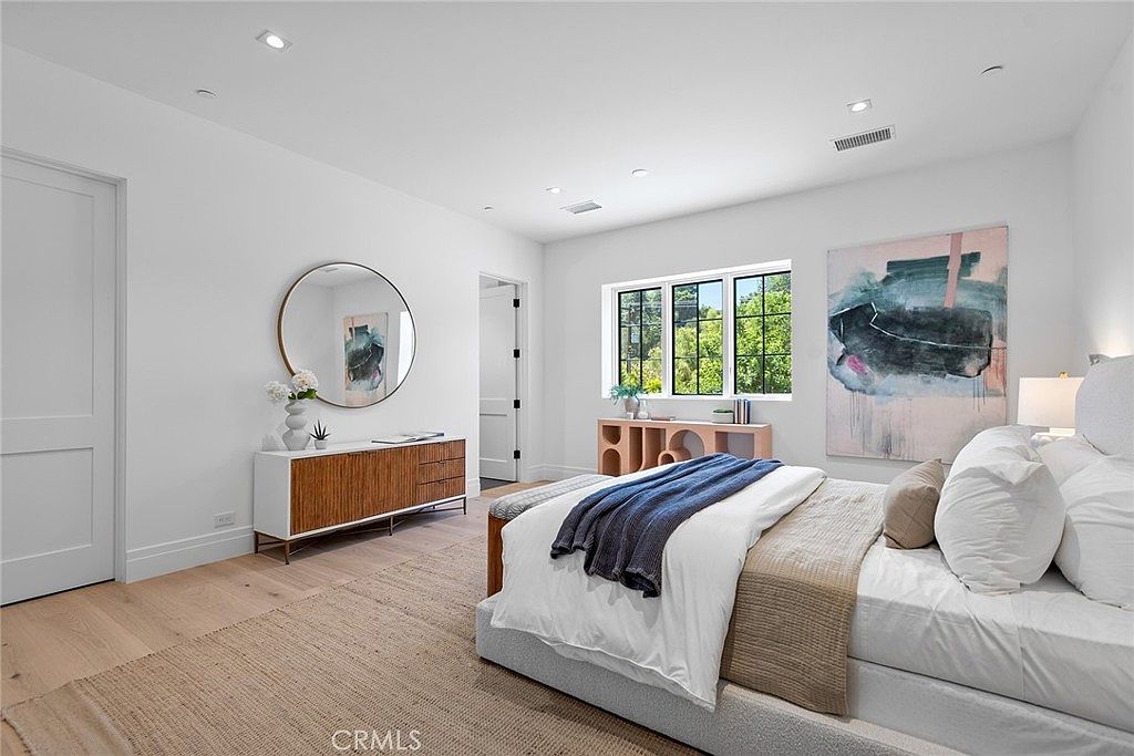 This bright and airy primary bedroom features light hardwood flooring, crisp white walls, and a large, plush bed with a navy blue throw blanket. A round mirror hangs above a modern wooden dresser, while a large abstract painting adds a pop of color to the space, creating a serene and sophisticated atmosphere. The room is well-lit by natural light streaming through the windows, highlighting the clean lines and contemporary design.
