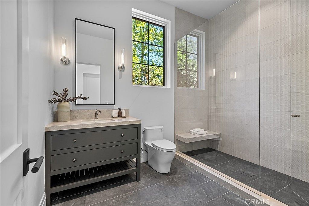This modern bathroom features a sleek dark gray vanity with a stone countertop, a rectangular mirror, and minimalist wall sconces. A spacious walk-in shower with a built-in stone bench and large-format tile walls sits adjacent to a white toilet. The room is brightened by a large window, creating a clean, contemporary, and functional aesthetic.