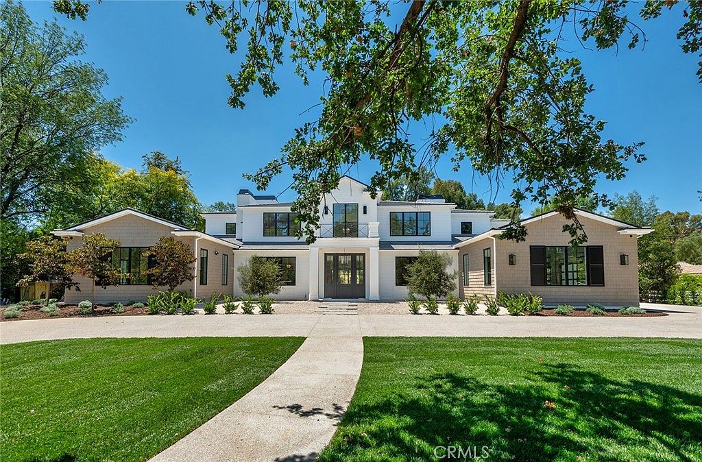 This stunning modern farmhouse features a symmetrical white facade with contrasting dark window frames and shingle-style siding on the wings. A wide, paved driveway leads directly to the grand central entryway, framed by lush green lawns and mature trees that provide a welcoming, upscale curb appeal. The perspective is a straight-on, eye-level shot that emphasizes the home's impressive scale and balanced architectural design.