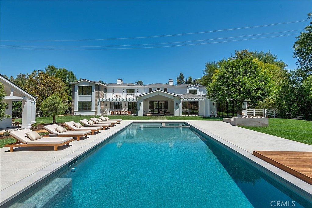 This stunning backyard features a long, rectangular swimming pool as the centerpiece, surrounded by a sleek stone deck and a row of modern lounge chairs. The expansive property includes a large, white multi-story residence with a covered patio area, lush green lawns, and mature trees, creating a serene and luxurious atmosphere. The perspective is captured from the edge of the pool, looking toward the house, emphasizing the seamless transition between indoor and outdoor living spaces.
