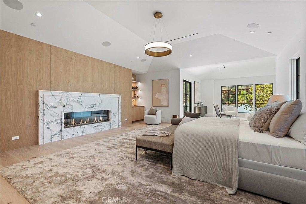 This spacious primary bedroom features a sophisticated, modern aesthetic with a large, plush area rug anchoring the room. A striking marble-clad fireplace serves as a focal point against a warm wood-paneled accent wall, while a contemporary chandelier hangs from the vaulted ceiling. The room is bathed in natural light from large windows, creating an airy and luxurious atmosphere.