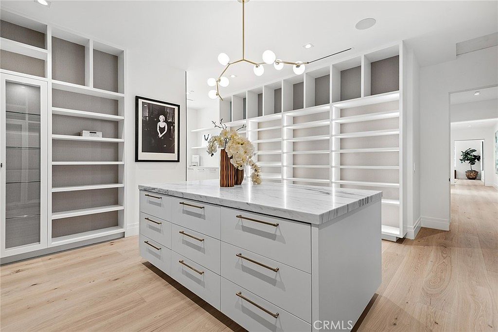 This luxurious walk-in closet features custom white built-in shelving and a central marble-topped island with ample drawer storage. A modern, multi-globe chandelier hangs above the island, while light wood flooring and a framed black-and-white photograph add a sophisticated, contemporary touch. The space is bright and airy, showcasing a high-end, organized design perfect for a primary suite.