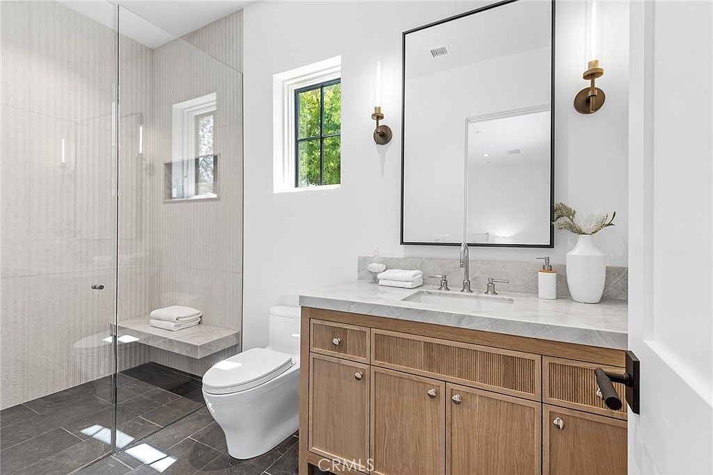 This modern, well-lit bathroom features a sleek vanity with a light wood finish, a marble countertop, and a rectangular undermount sink. To the left, a glass-enclosed walk-in shower with a built-in stone bench adds a luxurious touch, while a white toilet sits neatly between the shower and vanity. The space is accented by a large rectangular mirror, elegant wall-mounted light fixtures, and a neutral color palette that creates a clean, sophisticated atmosphere.