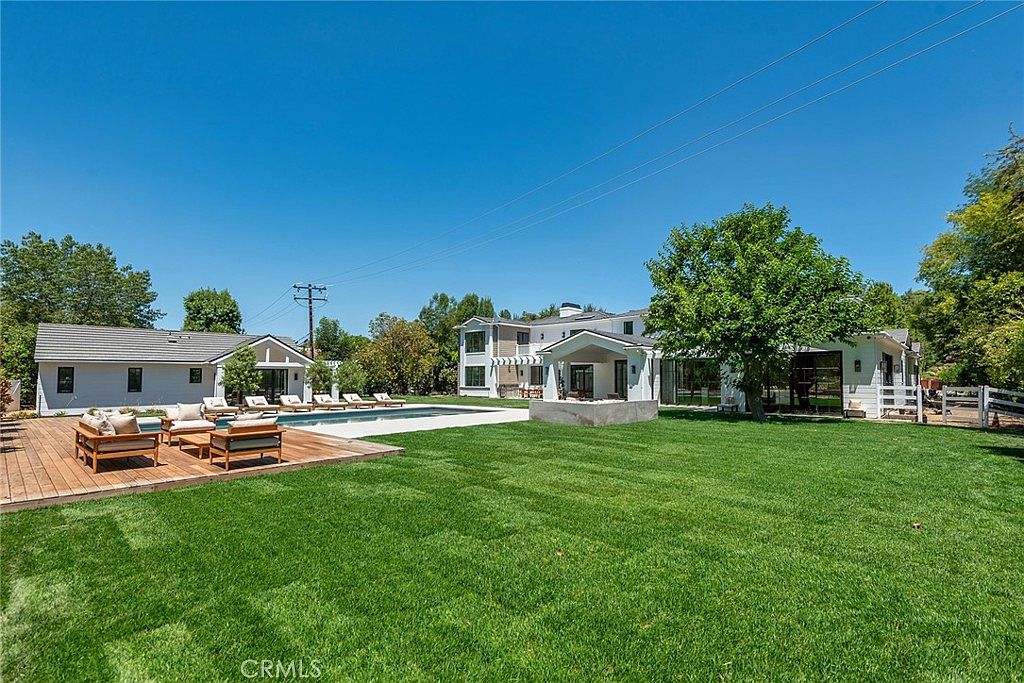 This expansive rear view captures a luxurious modern estate featuring a sprawling green lawn, a pristine swimming pool, and a wooden deck furnished with contemporary outdoor seating. The main residence and a detached structure showcase clean architectural lines, large glass doors, and a bright, airy aesthetic. The perspective is wide and inviting, emphasizing the seamless transition between indoor and outdoor living spaces in a sunny, well-manicured setting.