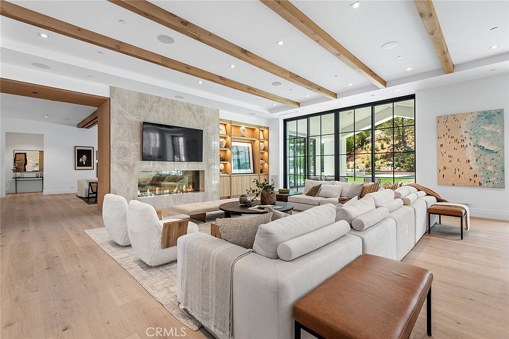This spacious, modern living room features a high ceiling with exposed wooden beams and a large, floor-to-ceiling marble fireplace as the central focal point. A massive, plush sectional sofa and two contemporary armchairs are arranged on a textured area rug, creating an inviting and luxurious seating area. Large glass sliding doors provide a seamless transition to the outdoors, while a piece of abstract art adds a vibrant touch to the neutral, sophisticated color palette.