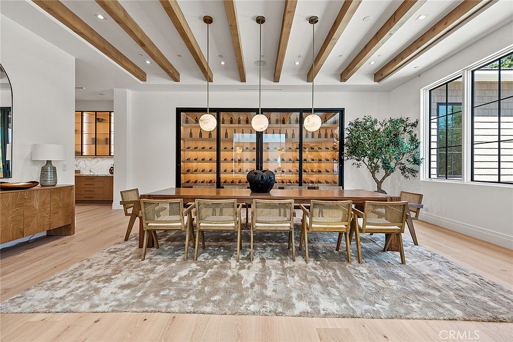 This elegant dining room features a long wooden table surrounded by eight woven-back chairs, set atop a large, textured area rug. The space is anchored by a striking floor-to-ceiling glass-enclosed wine display and illuminated by three modern spherical pendant lights hanging from a beamed ceiling. The room exudes a sophisticated, airy atmosphere with its neutral color palette and clean, contemporary design.