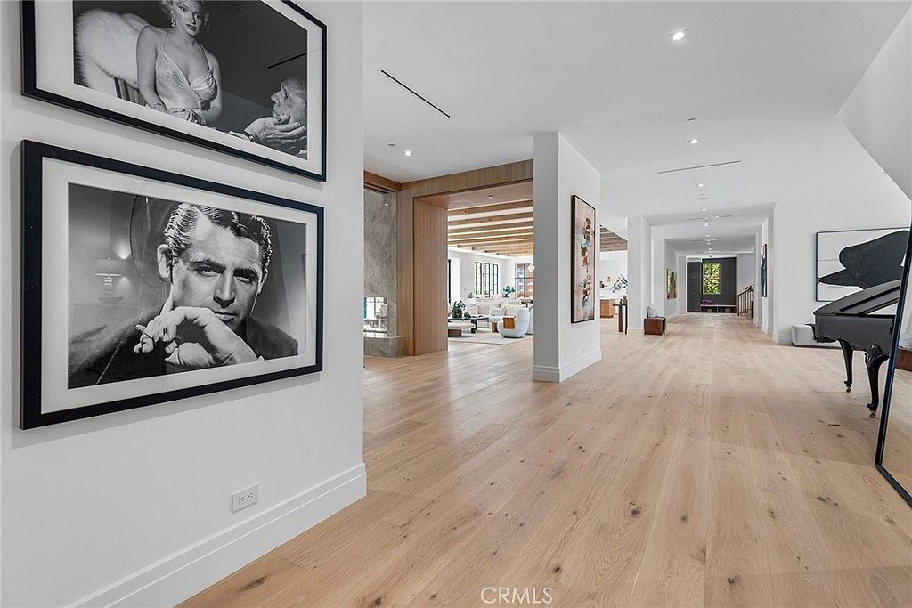 This expansive, light-filled hallway showcases a minimalist, modern aesthetic with light-toned hardwood flooring and crisp white walls. The space is adorned with large, framed black-and-white celebrity portraits, leading the eye toward a bright, open living area in the distance. A grand piano sits to the right, adding a touch of sophistication to the open-concept floor plan.
