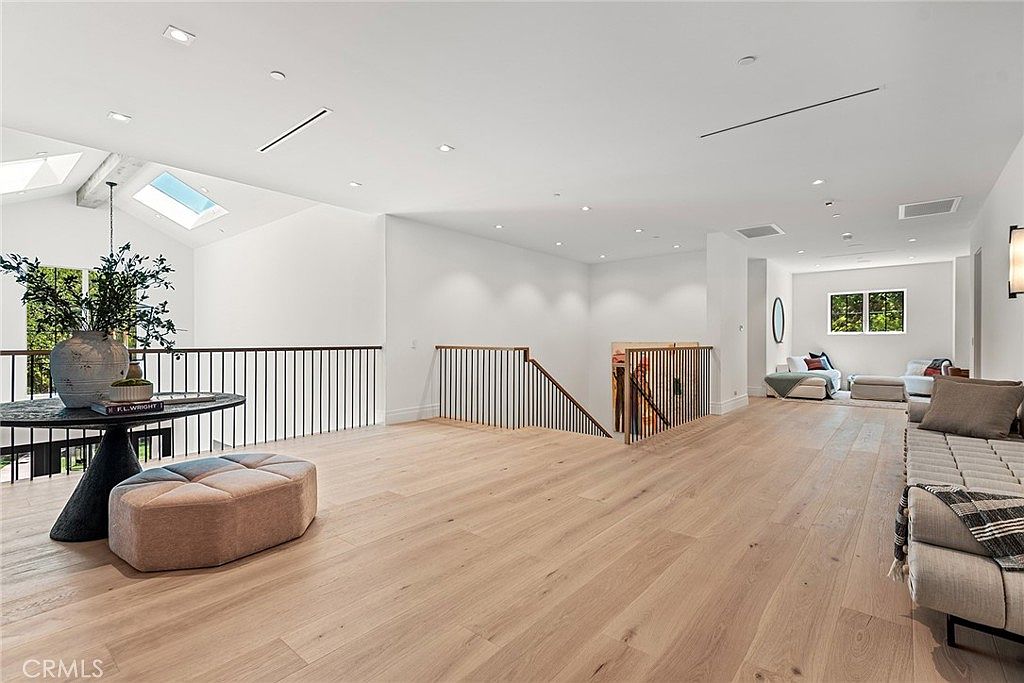 This spacious, light-filled landing area features high vaulted ceilings with skylights and sleek, light-toned hardwood flooring. A modern staircase with a minimalist black metal railing leads to the lower level, while the open floor plan connects to a comfortable seating area in the background. The aesthetic is clean, contemporary, and airy, emphasizing a sense of luxury and architectural openness.