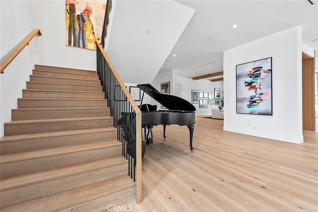 This elegant interior space features a grand staircase with light wood treads and a sleek black metal railing, serving as a focal point of the home. A polished black grand piano sits gracefully beneath the stairs, complemented by vibrant, modern abstract artwork on the walls. The open-concept design flows into a bright living area, showcasing light hardwood floors and a sophisticated, contemporary aesthetic.
