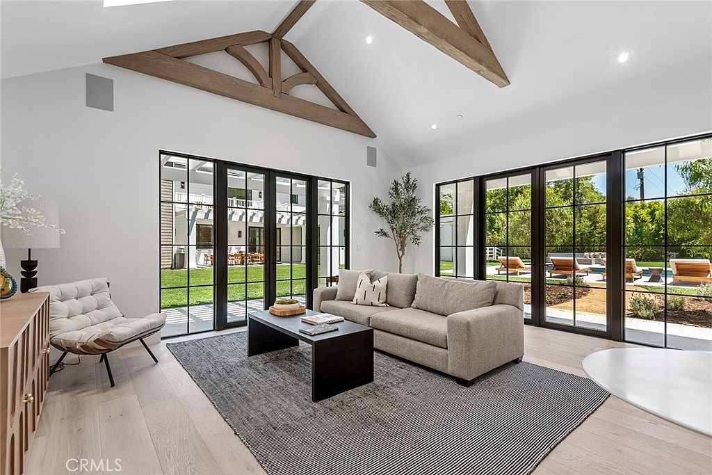 This bright and airy living room features a high vaulted ceiling with exposed wooden beams, creating a spacious and sophisticated atmosphere. Large floor-to-ceiling black-framed glass doors provide an abundance of natural light and a seamless transition to the outdoor patio and yard. The room is furnished with a neutral-toned sofa, a dark coffee table, and a textured area rug, offering a modern and inviting aesthetic.
