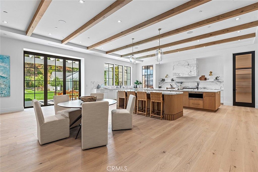 This expansive, open-concept kitchen and dining area features a sophisticated blend of modern design and warm, natural textures. A large central island with woven bar stools serves as the focal point, complemented by marble countertops, light wood flooring, and striking exposed wooden ceiling beams. The space is flooded with natural light from large windows and glass doors that provide a seamless transition to the outdoor patio, creating an airy and inviting atmosphere.