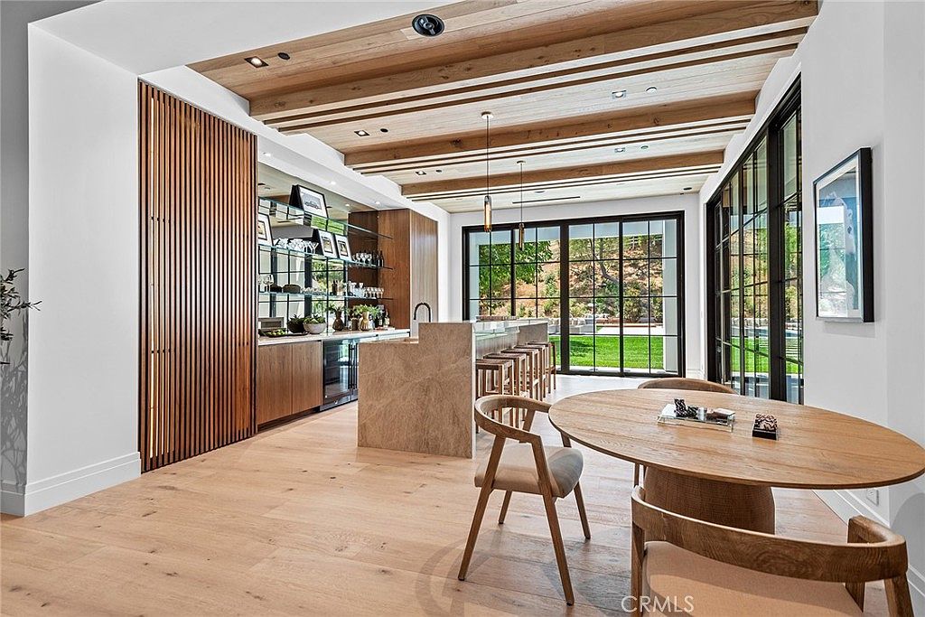 This sophisticated, open-concept kitchen and dining area features a stunning blend of natural wood textures, including a slatted wall and exposed ceiling beams. A large stone-topped island serves as a focal point, complemented by a sleek bar area with glass shelving and a wine fridge. The space is bathed in natural light from floor-to-ceiling black-framed windows that offer a seamless transition to the outdoor landscape.