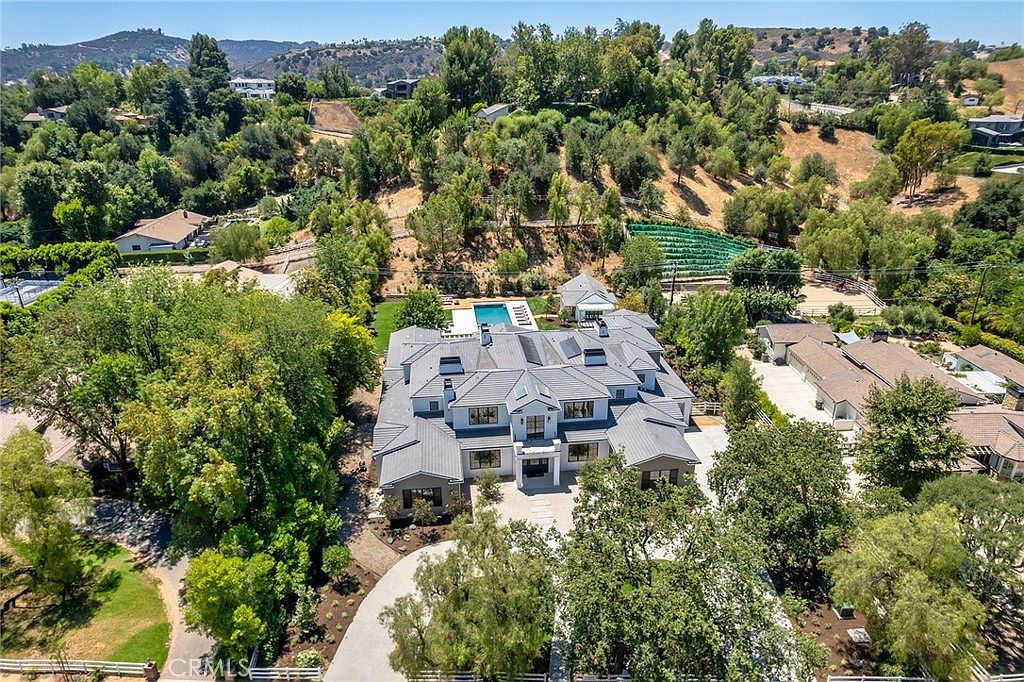 This high-angle aerial shot captures a sprawling, modern luxury estate nestled within a lush, hilly landscape. The property features a large multi-gabled roofline, a private swimming pool, and extensive surrounding greenery, showcasing a sense of seclusion and grandeur. The perspective emphasizes the expansive grounds and the architectural scale of the residence in its natural, serene environment.