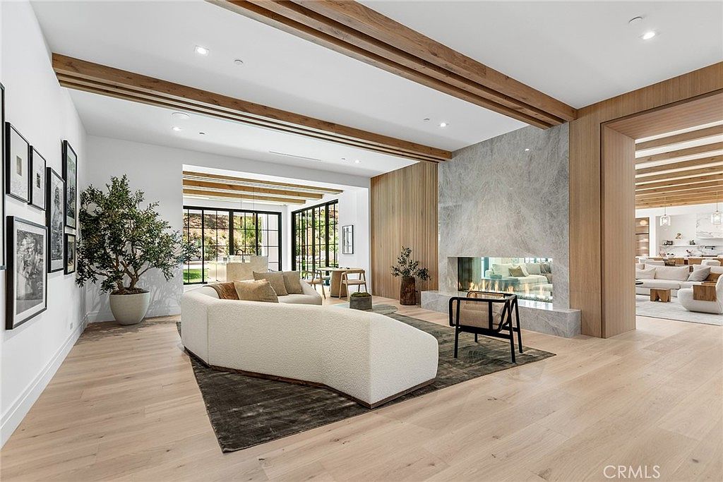 This expansive, open-concept living room features a sophisticated modern design with light hardwood flooring, a large curved cream sofa, and a striking floor-to-ceiling marble fireplace. The space is accented by warm wood ceiling beams and a gallery wall, creating a seamless flow into the adjacent dining area. The perspective is wide and inviting, showcasing a bright, airy atmosphere perfect for luxury living.