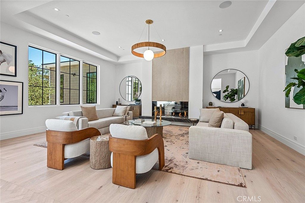 This sophisticated living room features a bright, open-concept design with light hardwood floors and a striking floor-to-ceiling fireplace as the focal point. The space is furnished with plush, neutral-toned sofas and modern wooden accent chairs, complemented by a large circular mirror and a contemporary pendant light. Large windows flood the room with natural light, creating an airy and inviting atmosphere perfect for high-end residential living.
