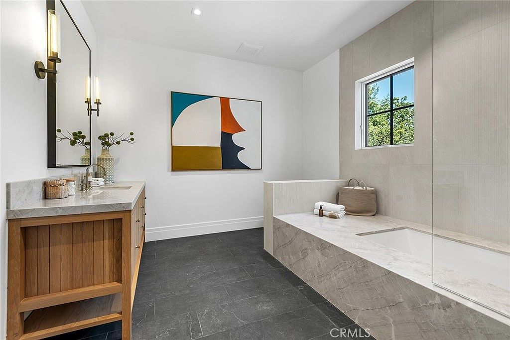 This luxurious primary bathroom features a sleek, modern design with a large soaking tub encased in elegant marble, complemented by a matching marble-topped vanity with a wooden base. A striking abstract painting adds a pop of color to the neutral, minimalist space, while a large mirror and contemporary lighting fixtures enhance the bright, airy atmosphere. The room is finished with dark, textured floor tiles that provide a sophisticated contrast to the light walls and stone surfaces.