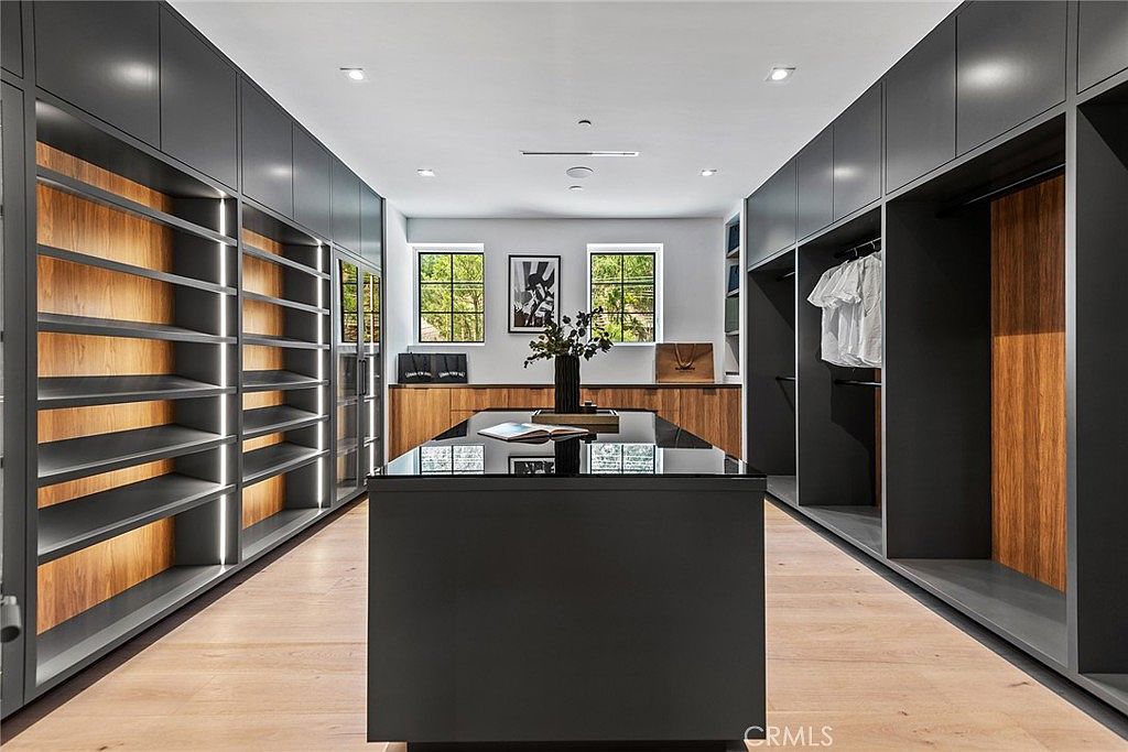 This luxurious, expansive walk-in closet features custom dark cabinetry with integrated warm LED lighting, providing a sophisticated and modern aesthetic. A central island with a sleek black countertop serves as a focal point, while light wood flooring adds warmth to the space. The symmetrical layout and clean lines create an organized, high-end boutique atmosphere.