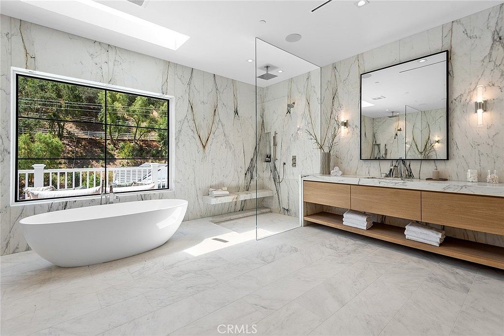 This luxurious primary bathroom features floor-to-ceiling marble tiling, a sleek freestanding soaking tub, and a spacious walk-in glass shower. A floating wooden vanity with integrated storage and a large, modern mirror adds warmth to the sophisticated, high-end design. The room is bathed in natural light from a large window, creating an airy and serene spa-like atmosphere.