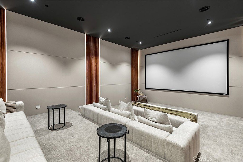 This image showcases a luxurious, tiered home theater room designed for an immersive cinematic experience. The space features a large projection screen, plush, light-colored sectional seating arranged on multiple levels, and sophisticated wall paneling with integrated wood accents. The dark ceiling and recessed lighting create a moody, professional atmosphere perfect for movie viewing.
